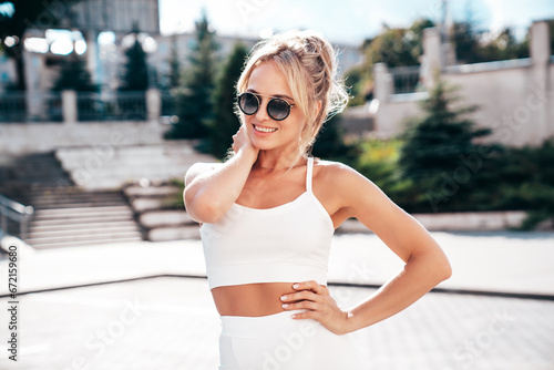 Young beautiful smiling female in trendy summer white cycling shorts and tank top clothes. Sexy woman posing in street. Positive model having fun. Cheerful and happy. At sunny day. In sunglasses