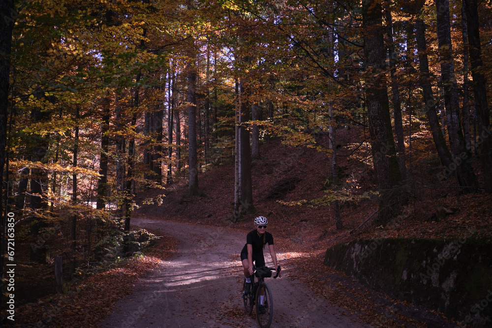 Fit female cyclist riding a gravel bike in autumn forest. Amazing autumn foliage. Cyclist training on beautiful autumn forest trail. Sport motivation. Adventure travel on bike. 