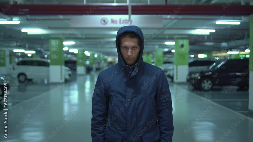 Hostile suspicious hooded young man stands and looks at camera in the underground parking Hostile suspicious hooded young man stands and looks at camera in the underground parking
