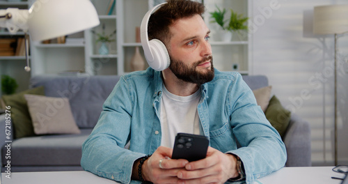 Young calm man 25s wear shirt headphones, sit for table hold and use mobile cell phone look aside listen music stay at home rest relax spend free spare time in living room, indoors © serg