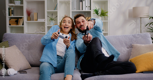 Happy couple sitting on the sofa playing video games, using controllers. competitive Girlfriend and boyfriend in love have fun playing in online video games in living room at home together