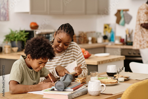 Smiling Black woman helping preteen son with doing homework for upcoming class