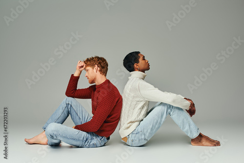 two young multiethnic men in cozy warm sweaters sitting on floor back to back, fashion concept