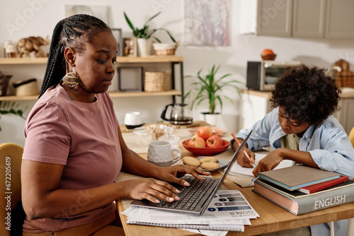 Black woman working on laptop at home when her son doing homework