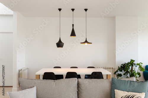 Front view of living room with white wooden table and six black chairs with three black metal lamps hanging from the ceiling, a plant and natural light. 