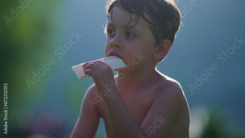 Pensive child eating biscuit while drying in the sun after playing at pool. Shirtless small boy enjoys summer vacations observing surroundings