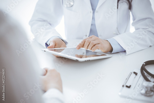 Doctor and patient sitting opposite each other at the desk in clinic. The focus is on female physician s hands using tablet computer  close up. Medicine concept