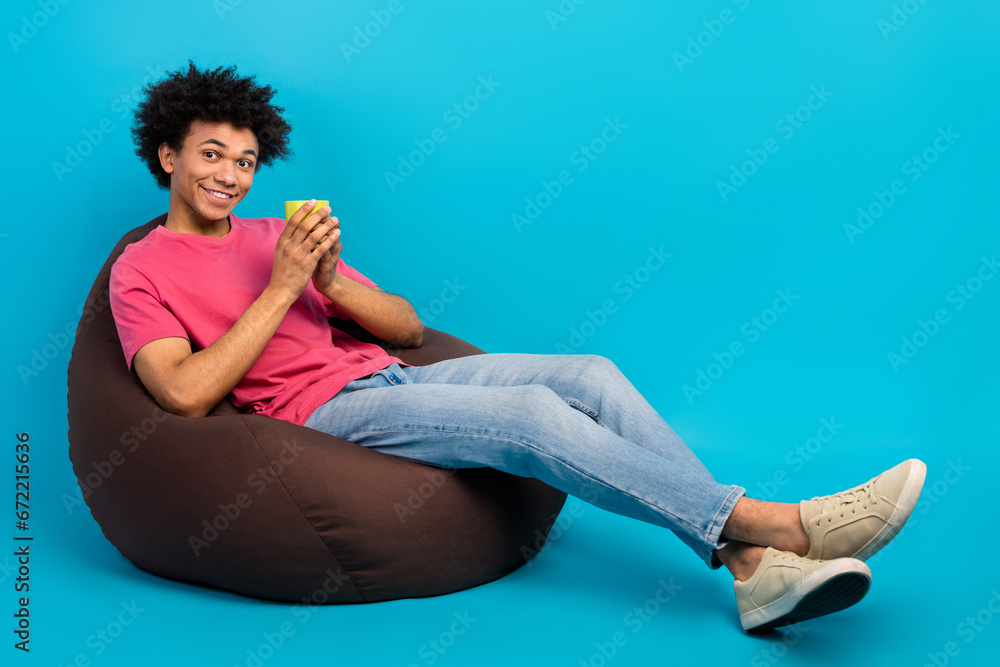 Full body photo of handsome young male sit brown beanbag hold coffee mug dressed stylish pink clothes isolated on blue color background Full body photo of handsome young male sit brown beanbag hold coffee mug dressed stylish pink clothes isolated on blue color background