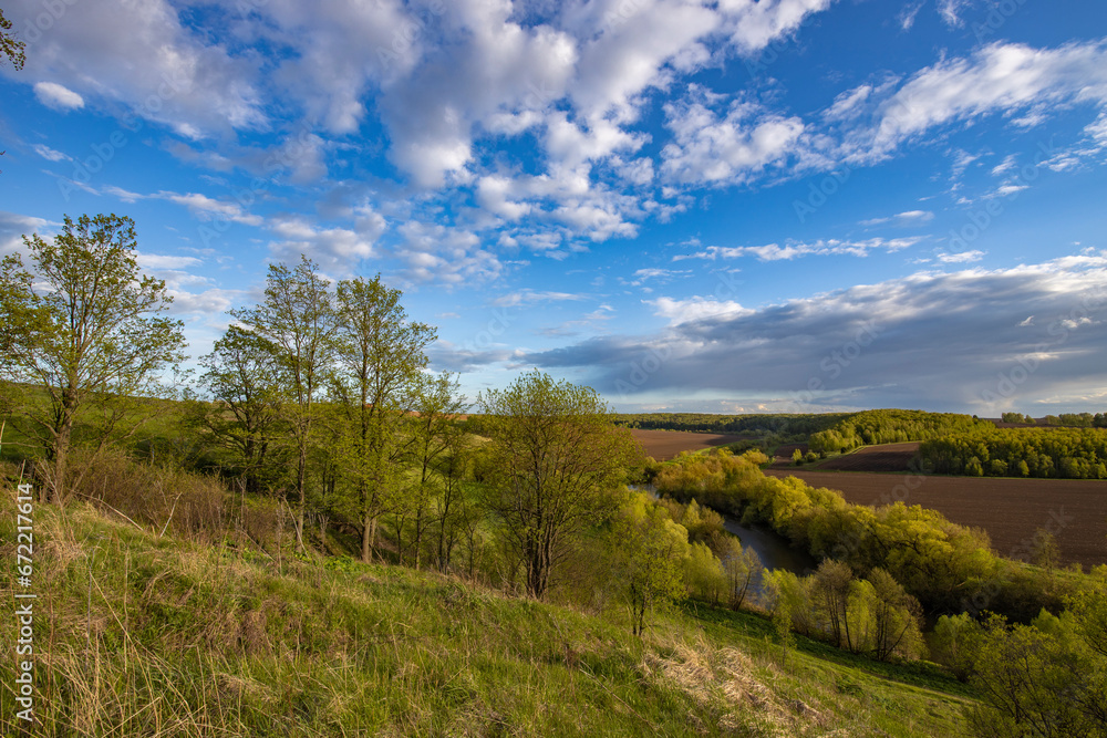 Fototapeta premium View from the hill to plowed farmland. Landscape with a river, arable land and green trees.
