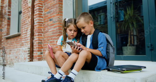 Two school little students small boy and girl sitting on stairs near modern school building. Pretty friends searching internet on smartphone and showing to friend. Concept of learning and lifestyle in