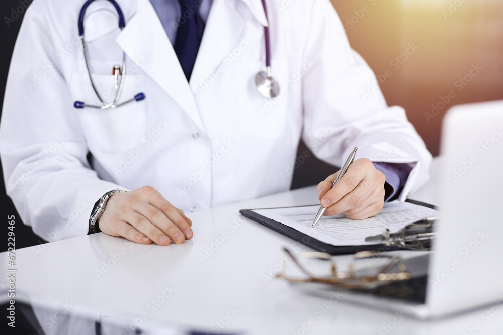 Unknown male doctor sitting and working with clipboard of medication history record in a darkened clinic, glare of light on the background, close-up of hands