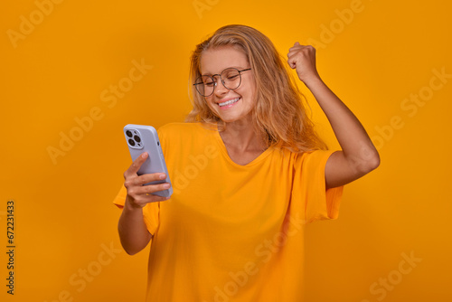 Young cheerful energetic Caucasian woman with phone happily waves hand and smiles celebrating victory in financial lottery and looks forward to receiving cash prize stands on yellow background.