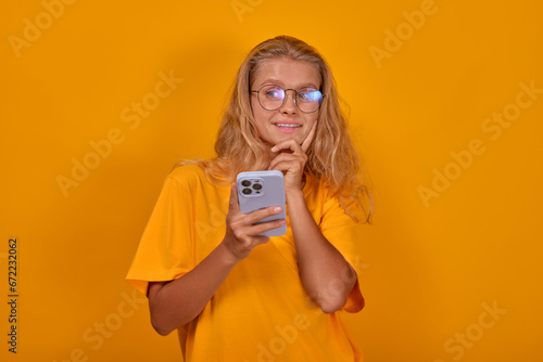 Young beautiful dreaming Caucasian woman teenager in glasses holding mobile phone coming up with new post for social network or idea for photo dressed in casual clothes stands in yellow studio.