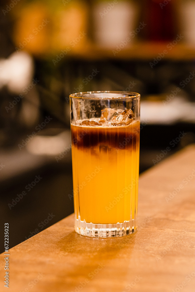 Bumble coffee with ice on a bar. Espresso, orange juice, syrup in layers in glass with ice. Refreshing coffee drink. Coffee house menu, recipe Bumble coffee with ice on a bar. Espresso, orange juice, syrup in layers in glass with ice. Refreshing coffee drink. Coffee house menu, recipe