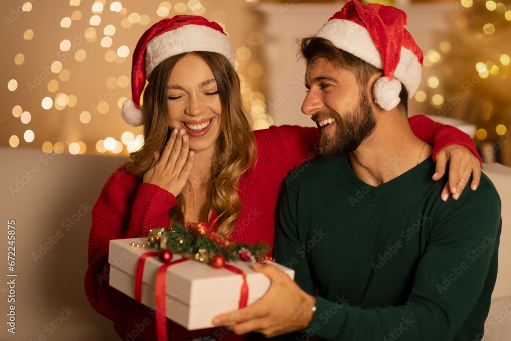 Romance and winter holidays. Surprised woman looks at gift box from husband, sitting in living room interior with Xmas tree with luminous garlands Romance and winter holidays. Surprised woman looks at gift box from husband, sitting in living room interior with Xmas tree with luminous garlands