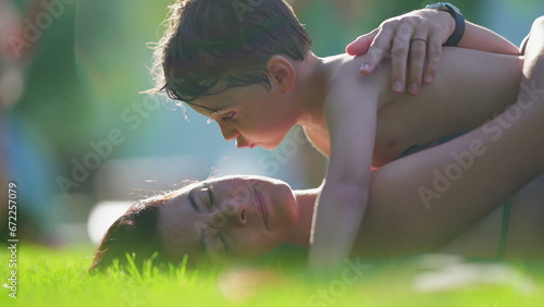 Affectionate moment of little son kissing mother in cheek while laid on grass during summer day vacations. Family lifestyle depicting love and care, skin-to-skin photo