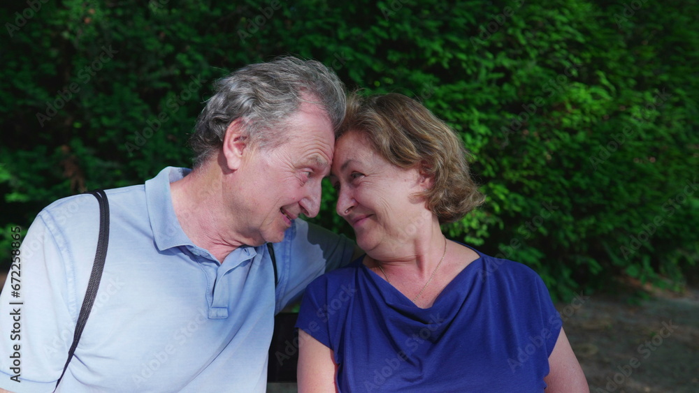 Funny moment between a senior couple sitting at park bench smiling and laughing while posing for camera during summer day Funny moment between a senior couple sitting at park bench smiling and laughing while posing for camera during summer day