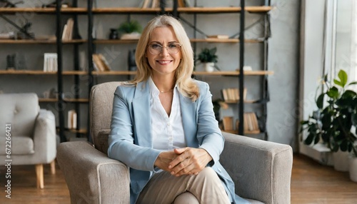 Middle aged female psychotherapist, counselor sitting in chair alone in office looking at camera. Sophisticated elegant mature 50s woman of mid age with blond hair posing indoors photo