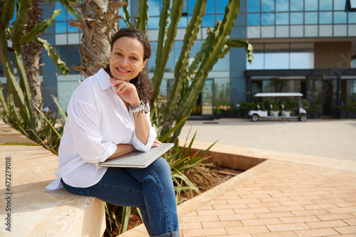 Beautiful woman in casual clothes, sitting on a stone bench with laptop on her knees, smiling cutely looking at camera