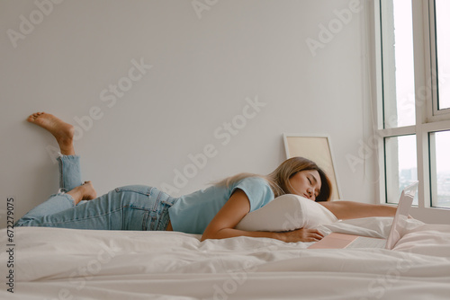 Asian Thai woman asleep on white bed while resting head on arm, using laptop for working at apartment room, tired sleeping of studying online by computer.