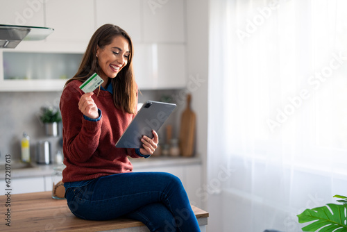 Happy young businesswoman holding credit card and looking at digital tablet pc while sitting on kitchen counter at home.