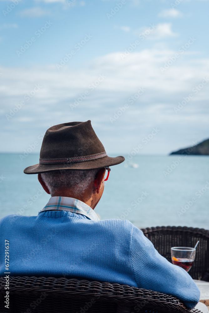 Homme avec un verre regardant la mer. Homme assis en terrasse sur la plage. Se relaxer à la mer. Prendre des vacances. Retraité dans le Sud. Sénior en vacances. Apéritif à la mer. Regarder l'horizon