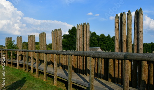 A close up on an old borough or village surrounded by a wooden palisade made out of logs, planks, and boards and a small moat below the hill seen on a sunny summer day next to some forests and moors