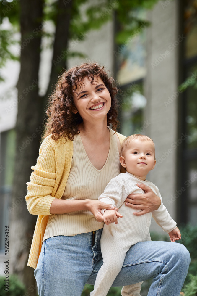 portrait of smiling and curly woman with cute toddler daughter on urban street, happy motherhood portrait of smiling and curly woman with cute toddler daughter on urban street, happy motherhood