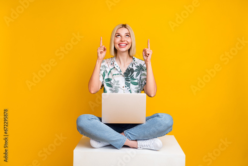 Full length photo of clever pretty girl look directing at sale empty space sit on cube with laptop isolated on yellow color background © deagreez