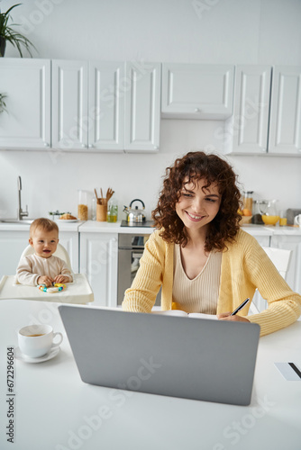 smiling woman working on laptop near coffee cup and toddler daughter on background in kitchen