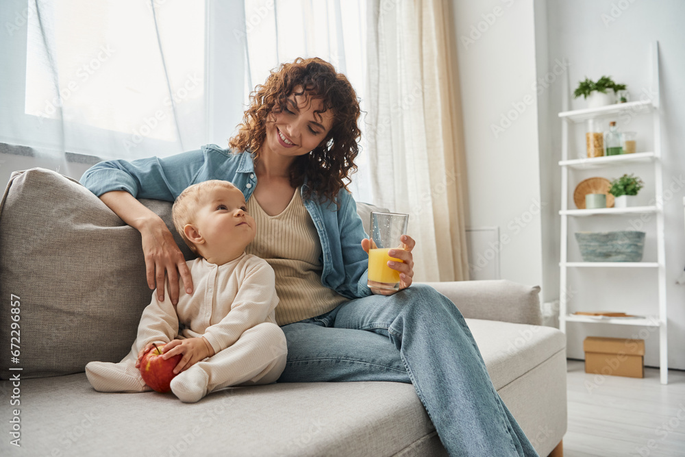 mother and toddler girl with orange juice and fresh apple looking at each other on cozy couch mother and toddler girl with orange juice and fresh apple looking at each other on cozy couch