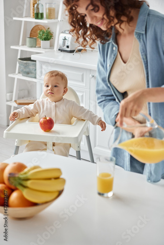 cute child sitting in baby chair near apple while happy mom pouring fresh orange juice for breakfast