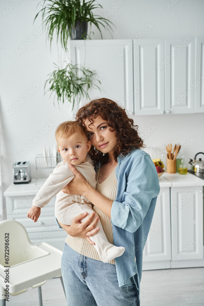 mother looking at camera while holding cute little daughter in modern kitchen at home, love and care mother looking at camera while holding cute little daughter in modern kitchen at home, love and care