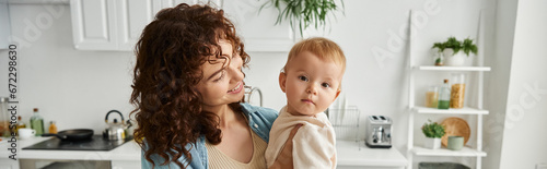 Wallpaper Mural woman with wavy hair smiling near cute baby girl in kitchen, happy motherhood, horizontal banner Torontodigital.ca