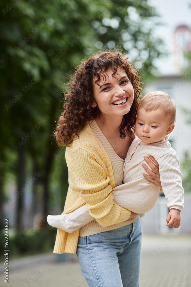Fototapeta premium curly woman smiling at camera while holding little daughter on urban street, happy modern family