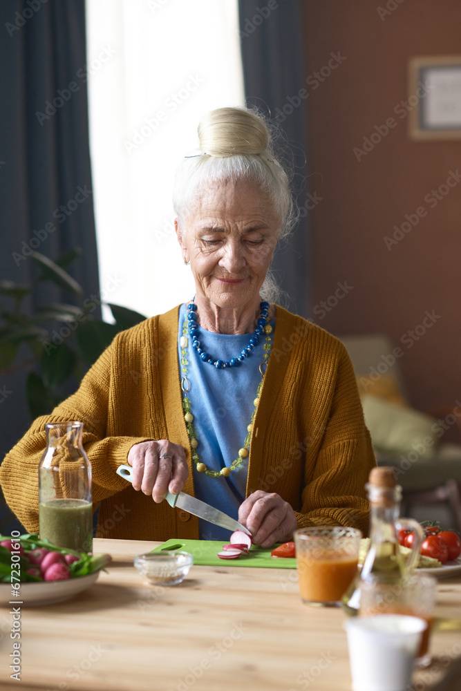 Vertical image of senior woman cutting vegetables for salad while sitting at table in the room Vertical image of senior woman cutting vegetables for salad while sitting at table in the room