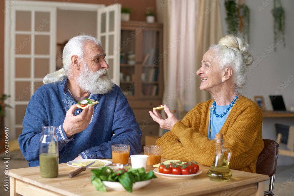 Senior couple eating sandwiches and talking to each other while having breakfast at home Senior couple eating sandwiches and talking to each other while having breakfast at home