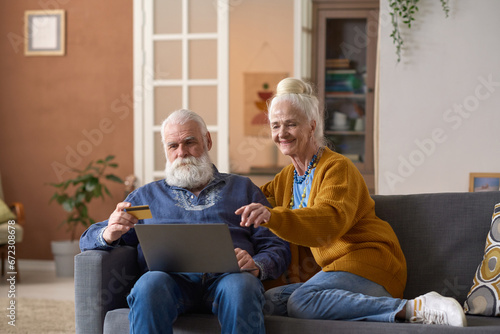 Senior couple sitting on sofa with laptop in the living room and using credit card to pay for purchase online