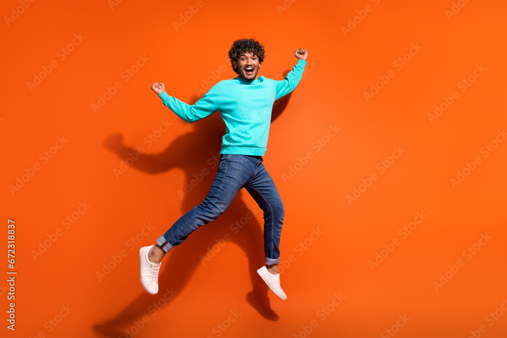 Full length photo of sportive indian guy curls hair wear denim jeans with cyan pullover win lottery isolated on orange color background