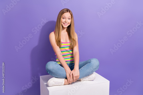 Full length photo of lovely teen blonde lady folded legs sit white cube wear trendy striped garment isolated on purple color background