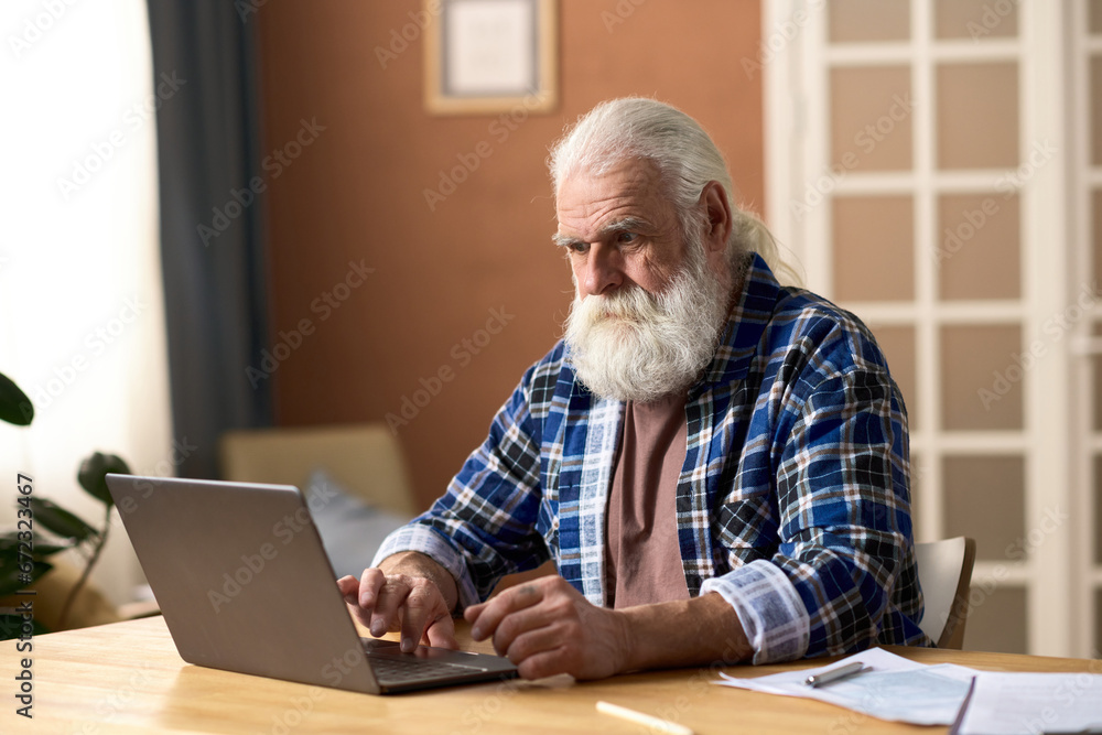Serious senior man typing on laptop while sitting at table in the room Serious senior man typing on laptop while sitting at table in the room