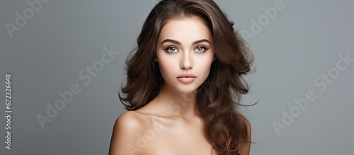 A stunning young woman with brown hair styled back a clear and radiant complexion large eyes and exposed shoulders striking a pose against a gray studio backdrop in a beautiful photograph ap