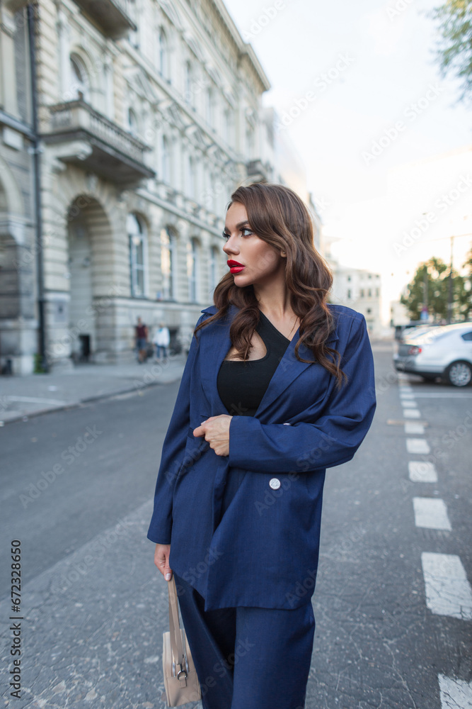 Beautiful fashion young business lady with a stylish blue dress in a fashionable blue suit walks on the street near a vintage building