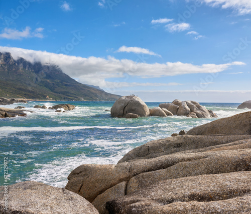 A calming ocean view of the rocky coast of Camps Bay in Cape Town, South Africa. Boulders at a beach with the tide and current coming in and Table Mountain on the horizon. Tidal waves washing ashore