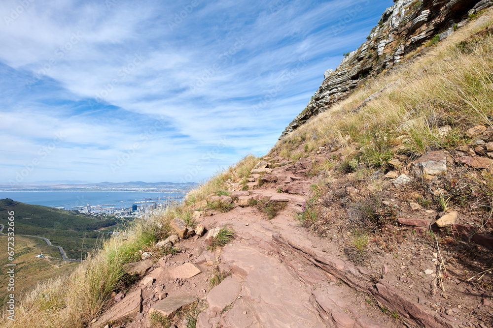 Scenic of a rocky mountain slope with view of a coastal city by the sea and cloudy sky background with copyspace. Rugged landscape on a cliff with hiking trail on Lions Head, Cape Town, South Africa Scenic of a rocky mountain slope with view of a coastal city by the sea and cloudy sky background with copyspace. Rugged landscape on a cliff with hiking trail on Lions Head, Cape Town, South Africa