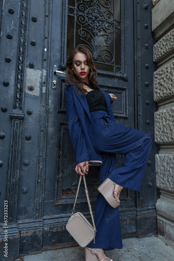 Pretty elegant fashion chic woman with hairstyle and red lips in fashionable blue suit with stylish bag and shoes stands and poses near a vintage black door on the street. Beautiful lady