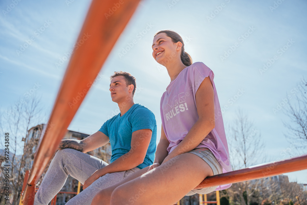 Sports friends taking a break from workout. They are sitting on horizontal metal bars in the park and looking away. Low angle view shot
