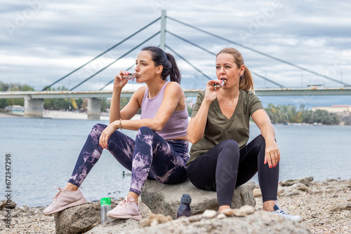 Two female friends pursuing their activity outdoors. Resting by the river bank after jogging, and refreshing with a healthy snack or a cereal bar.