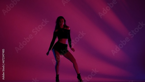 Attractive woman dancing heels dance in a studio. Pink and blue neon lights, striped shadowed background. Black sexy costume, high heels. Modern sensual choreography. Full length.