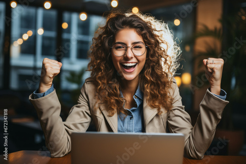 Joyful woman with curly hair celebrating success at a laptop in a cafe photo